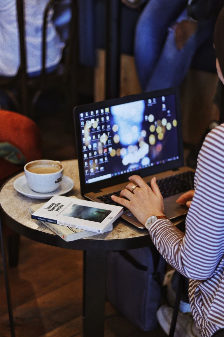Crop anonymous female remote employee working on netbook at cafeteria table with books and cappuccino against people